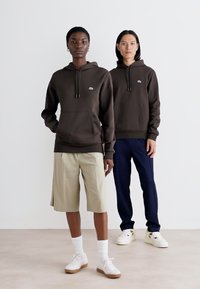 Sudadera con capucha marrón y bolsillo canguro, llevada por dos modelos; uno con pantalones cortos beige y el otro con pantalones azul marino y zapatillas blancas.