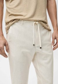 Jasnobeżowe spodnie na sznurek z pionowymi paskami, wykonane z teksturowanej tkaniny. Posiadają kontrastowe wiązania z metalowymi elementami.