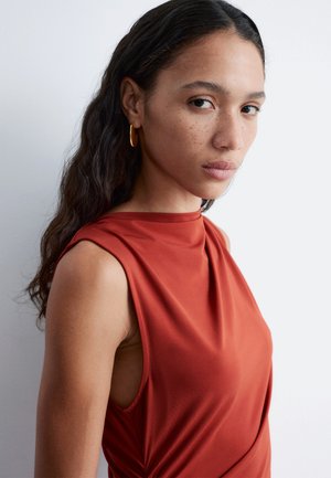 Donna con capelli lunghi e mossi, che indossa un top senza maniche color ruggine e orecchini a cerchio dorati, che guarda verso la fotocamera con un'espressione neutra.