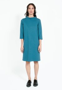 Teal knälång klänning med trekvartsärmar, en cowl-hals och en avslappnad passform. Bärs med svarta platåskor. Slätt, mjukt tyg.