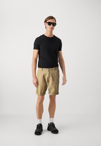 Camiseta negra de manga corta, pantalones cortos beige enrollados y zapatillas negras con calcetines blancos. El modelo lleva gafas de sol negras y se encuentra sobre un fondo neutro.