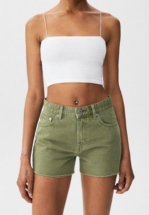 Femme portant un haut court blanc à fines bretelles et un short en jean vert olive taille haute avec un ourlet effiloché et un piercing au nombril.