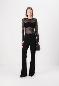 Crna čipkasta bluza s dugim rukavima s čvrstim crnim remenom, uparena s crnim hlačama visokog struka. Outfit upotpunjuje mala crna torbica.