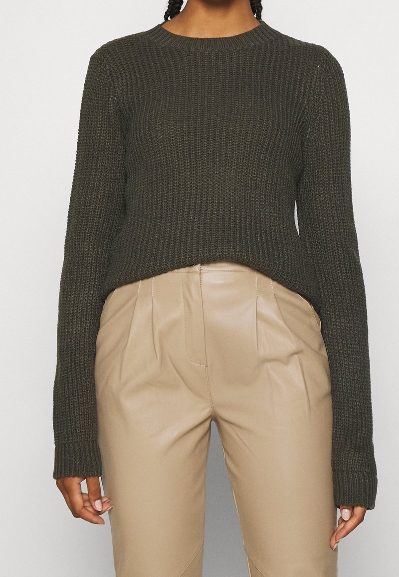 Pull en maille vert olive à manches longues, texture côtelée, et ourlet raccourci, associé à un pantalon beige sur mesure.