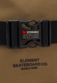 Bolsa de tela marrón con una superficie texturizada, que presenta correas ajustables negras y una hebilla de plástico negra. Logotipo de la marca visible en la parte frontal.