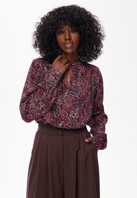 Vrouw met krullend haar, gekleed in een gebloemde blouse met lange mouwen en hoge bruine broeken, poseert met één hand die haar kraag aanraakt en de andere in haar zak.