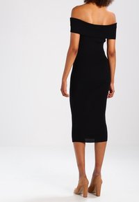 Robe midi noire, épaules dénudées, silhouette ajustée, texture lisse et ourlet au niveau des genoux. Portée avec des chaussures à talons hauts couleur nude.