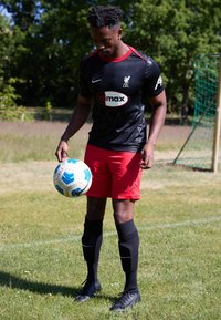Maglia da calcio nera con accenti rossi, pantaloncini rossi e calze nere; il giocatore tiene in mano un pallone da calcio blu e bianco su un campo erboso.