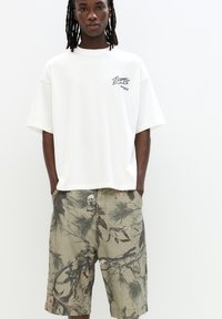 T-shirt bianco oversize con design grafico sul lato sinistro del petto, abbinato a pantaloni corti con motivo mimetico in tonalità verdi, marroni e nere.
