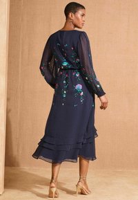 Rochie bleumarin cu volane, realizată dintr-un material ușor, cu broderie florală. Mâneci lungi, talie strânsă și detalii cu fuste stratificate.