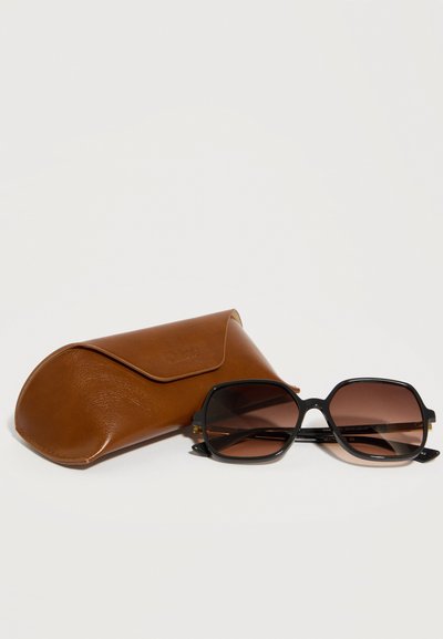 Μαύρα υπερμεγέθη τετράγωνα γυαλιά ηλίου με καφέ φακούς με βαθμιαία απόχρωση δίπλα σε καφέ δερμάτινη θήκη προστασίας σε λευκό φόντο.