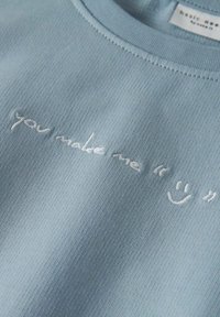 Camisa de manga corta azul claro con textura acanalada y cuello redondo. Presenta un texto bordado en blanco: "me haces sentir 😊."