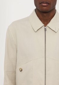 Man wearing a beige jacket with a pointed collar, front zipper, and button detail on the left side, shown from shoulders to mid-chest.