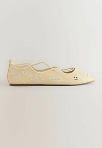 Chaussure plate couleur crème avec bout pointu, broderie florale blanche et fines lanières croisées à la cheville, vue de côté.