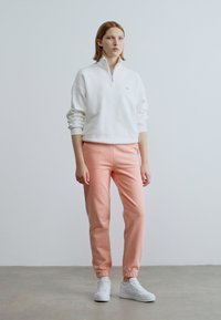 Sweatshirt branco com fecho de correr e calças de treino cor de rosa claro, com punhos elásticos e corte justo. O modelo usa ténis brancos, posando contra um fundo neutro.