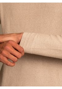 Bež kootud sviiter ribidega äärtega, näidates pehmet tekstuuri ja peeneid horisontaalseid jooni, mille hoiab käsi sobivas taustas.