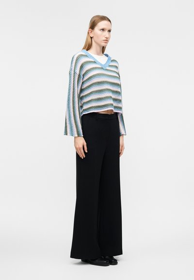 Femme debout portant un pull à col en V rayé bleu, violet et vert avec des manches larges et un pantalon noir à jambes larges, sur un fond uni.