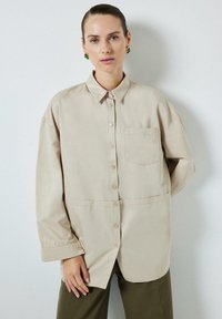 Beige, hosszú ujjú ing gombos gallérral, elülső zsebbel és puha tapintással, olajzöld nadrággal párosítva.