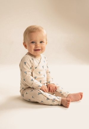 Usmievajúce sa dieťa so svetlými vlasmi sedí bosé na podlahe, oblečené v béžových pyžamách s modrými potlačami balónov na neutrálnom pozadí.