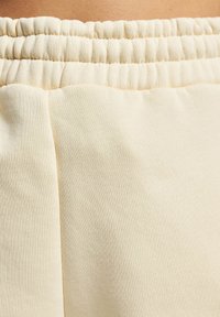 Tăblia din stofă bej deschis prezintă o textură moale, având un brâu strâns. Designul include un detaliu de cusătură vizibil, subliniind stilul vestimentar casual.