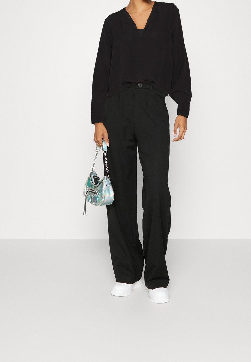 Personne portant un haut noir à manches longues et un pantalon à jambes larges tenant un petit sac à main argenté brillant, portant des chaussures blanches, debout sur un sol gris.