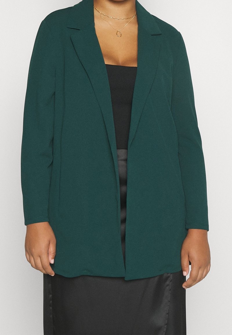 Femme portant un blazer vert foncé par-dessus un haut noir et une jupe en satin noire, accessoirisée avec des colliers dorés superposés.