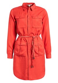 Robe-chemise rouge à manches longues avec patte de boutonnage à l’avant, quatre poches à rabat boutonnées, col et fine ceinture nouée à la taille.