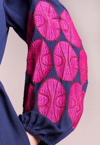 Mörkblå tygärm med livliga rosa broderade ovala mönster och vågiga kanter, med detaljerad manschett med rynkor.