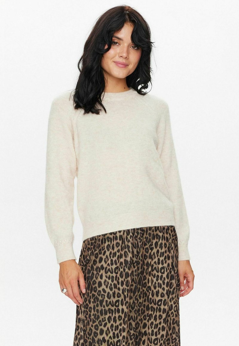 Svetlobežový pletený sveter s dlhými rukávmi a okrúhlym výstrihom, nosený s tmavými sukňami s leopardím vzorom, s jemnou textúrou a voľným strihom.