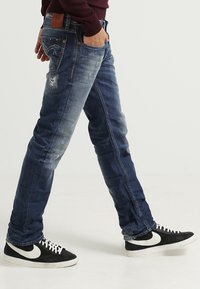 Jeans de mezclilla azul oscuro con un acabado desgastado, que presentan un corte ajustado y un desvanecimiento sutil. Combinados con zapatillas negras con acentos blancos.