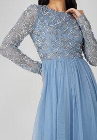 Vestido azul de manga larga con un cuerpo de encaje bordado, mangas transparentes y falda plisada y fluida. Detalles de cuentas plateadas en todo el cuerpo.