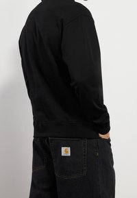 Sort sweatshirt med rund halsudskæring og ribbede ærmekanter, båret med mørke jeans med en logo-lap på baglommen.