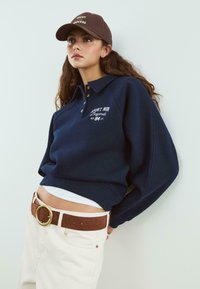Junge Frau, die einen marineblauen Pullover mit dem Text "Sport Mode Legends", beigen Hosen mit einem braunen Gürtel und einer braunen Kappe trägt, lehnt an einer Wand.