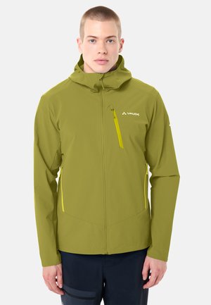 Junger Mann trägt eine olivgrüne Kapuzenjacke mit gelben Reißverschlusstaschen und dunkler Hose, steht vor einem schlichten hellen Hintergrund.