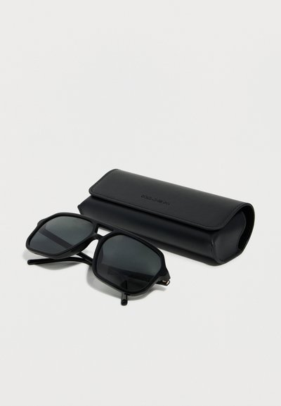 Μαύρα ορθογώνια γυαλιά ηλίου με σκουρα φακούς που ακουμπούν δίπλα σε μαύρη δερμάτινη θήκη με το λογότυπο Dolce & Gabbana πάνω σε λευκή επιφάνεια.