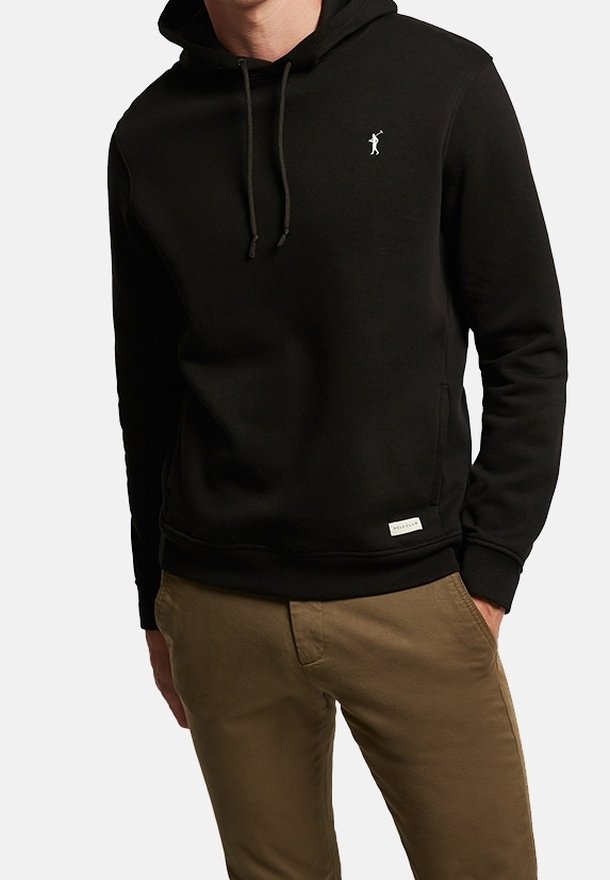 Personne portant un sweat à capuche noir avec un petit logo de golfeur blanc sur la poitrine et un pantalon marron, les mains dans les poches, sur un fond uni.