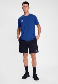 Camiseta azul de algodón con logo, pantalones cortos negros con logo, zapatillas deportivas negras, calcetines negros, de pie sobre un fondo blanco liso.