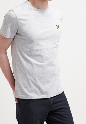 Uomo che indossa una maglietta a maniche corte grigio chiaro con un piccolo logo giallo sul petto e pantaloni scuri, in piedi con le mani dietro la schiena.