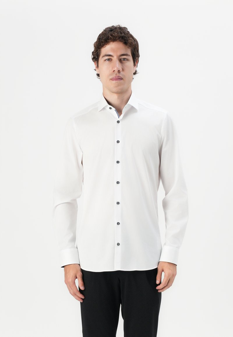 Chemise blanche à manches longues avec un col pointu et des boutons noirs sur le devant, présentant un léger éclat et des détails de poignets contrastés.