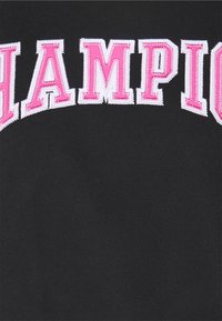 Svart tyg med stora "CHAMPION"-text i upphöjda rosa bokstäver, markerade med vit söm. Texturen är slät och lätt töjbar.