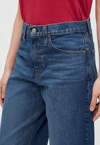 Personne portant un jean en denim bleu avec des poches avant et une chemise rouge rentrée, montrant la ceinture et le détail du bouton.