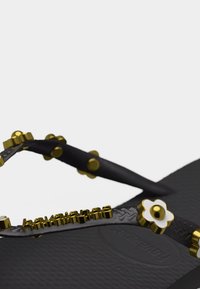 Μαύρο σανδάλι τύπου φλιπ-φλοπ με διακοσμημένες τιράντες που διαθέτουν χρυσά τρουκς και λευκές διακοσμήσεις σε σχήμα λουλουδιού, σε ανοιχτό γκρι φόντο.