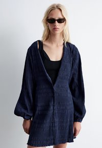 Marineblaues Hemdjacke mit Knöpfen aus strukturiertem Stoff, weiten Ärmeln und einem lockeren Schnitt. Über einem schwarzen gerippten Tanktop getragen.