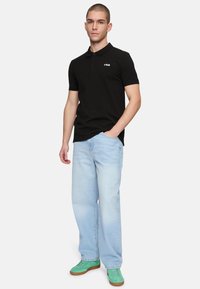 Svart pikétröja med krage och tre knappar, prydd med en liten vit logotyp. Kombinerad med ljusblå baggy-jeans och gröna sneakers.