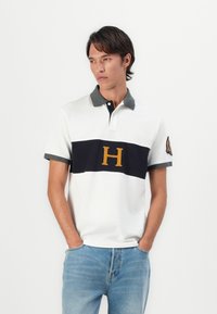 Valkoinen polo-paita, jossa on tummansiniset vaakaraidat ja suuri keltainen "H"-logo, harmaa kaulus ja hihojen suut, yhdistettynä vaaleansinisiin farkkuihin.