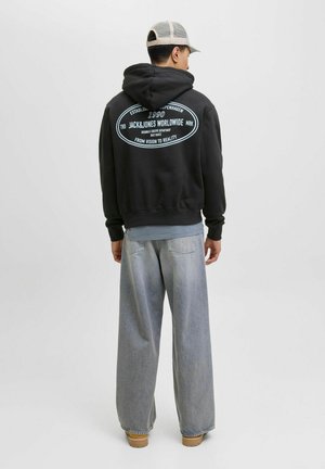 Schwarzer Kapuzenpullover mit großem Drucklogo auf dem Rücken, kombiniert mit hellblauen, weiten Jeanshosen und beigem Schuhwerk.