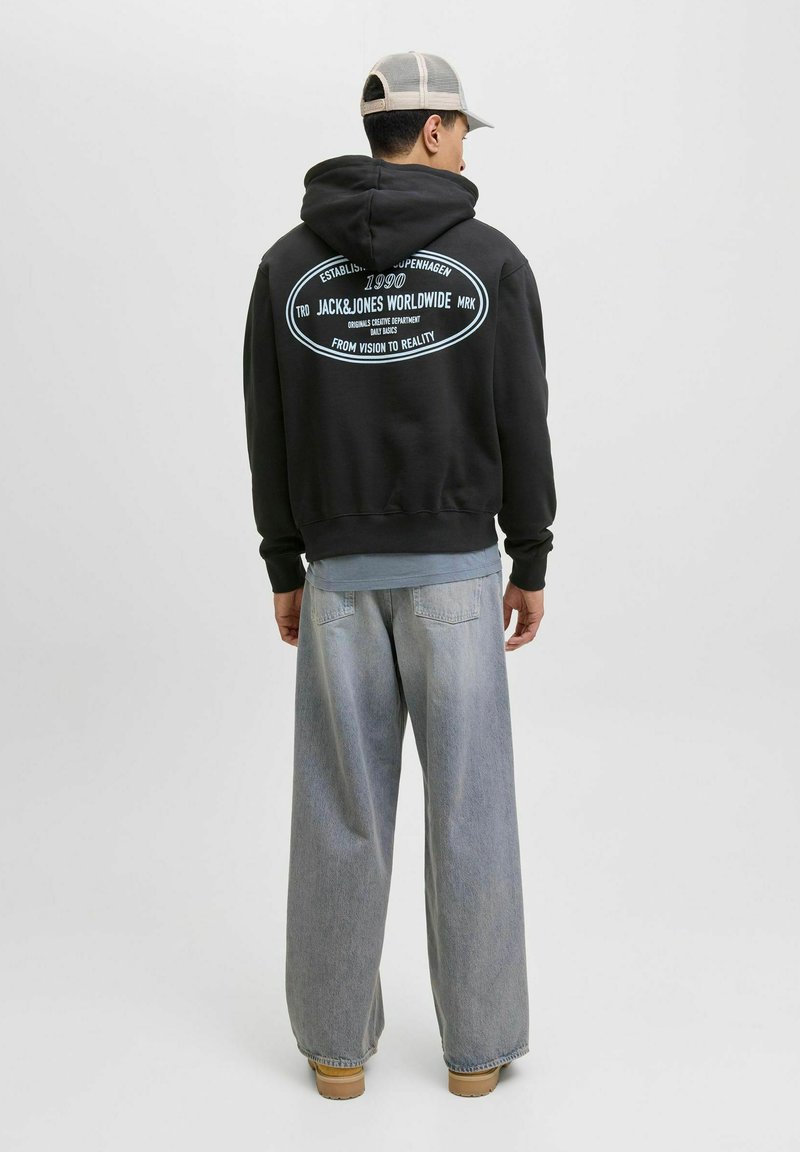 Sudadera negra con un gran logo impreso en la espalda, combinada con pantalones de mezclilla de tiro alto y pernera ancha, y calzado en tono marrón claro.