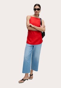 Haut rouge sans manches à coupe décontractée, associé à un jean en denim bleu clair à jambes larges. Le modèle porte des lunettes de soleil noires et des sandales.