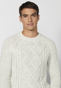 Cremfarbener Strickpullover mit Rundhalsausschnitt, der ein Rautenmuster aus Zopfstrick und gerippte Bündchen aufweist. Die Textur ist grob und strukturiert.