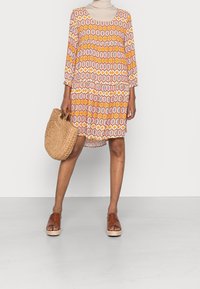 Patroondelige jurk met korte mouwen in oranje, crème en roze met een vierkante halslijn. Een geweven leren tas en bruine sleehakken maken de look compleet.